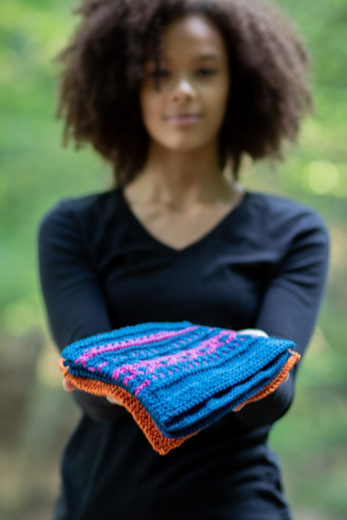 A model holds out a stack of beautiful knitting swatches as if to give them to the viewer